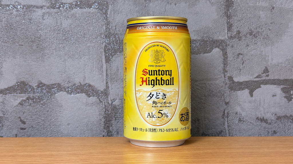 グレーの柄入り壁紙と木の天板を背景に置いた角ハイボール夕どきの缶